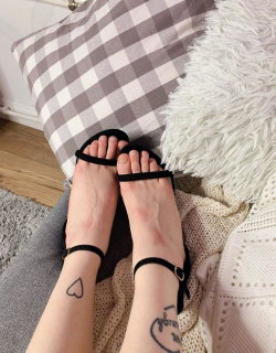 Person with tattoos on their legs wearing black sandals on a bed with a checkered pillow.