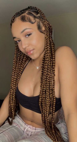 A woman with long braided hair wears a black crop top and light-colored pants, posing indoors in a selfie-style shot.