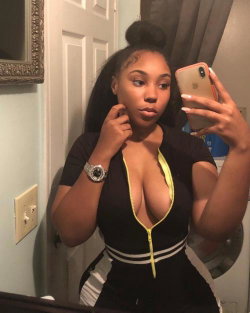 A curvy Black woman in a black zip-front top with yellow trim takes a mirror selfie, her hair styled in a high bun.
