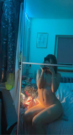 A woman with a slim build sits nude on a bed in a dimly lit room, taking a mirror selfie with blue ambient lighting.
