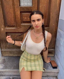 A young woman with braided hair wears a white tank top and plaid mini skirt, posing on stone steps in front of a wooden