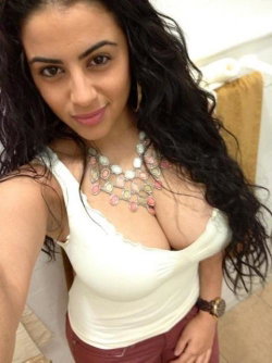 A woman with long dark hair wears a white tank top and red pants, smiling at the camera in a selfie-style pose indoors.