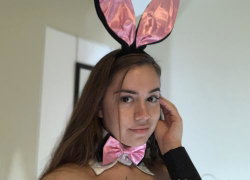 Brunette woman wearing pink bunny ears and bow tie, posing indoors with hand near face.