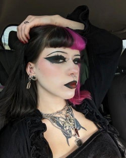 Woman with black and pink hair, dramatic makeup, multiple piercings, and a chest tattoo, wearing a black ruffled top
