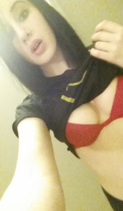 A woman with long dark hair takes a selfie, pulling her black shirt to reveal a red bra, in a dimly lit indoor setting.