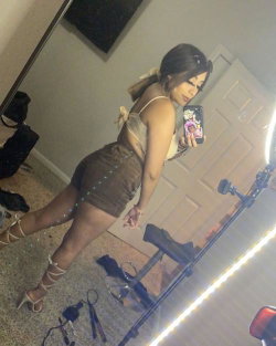 A woman with dark hair in a ponytail takes a mirror selfie wearing a beige crop top, brown shorts, and strappy heels in