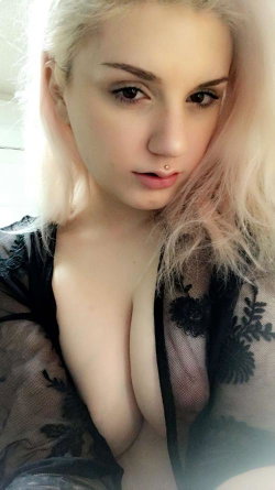 Blonde woman with light hair and a lip piercing, wearing a sheer black lace top, posing closely in a selfie-style shot.
