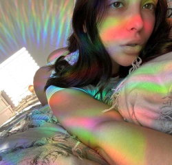 Brunette woman with wavy hair lying on a bed, wearing a white top, illuminated by colorful rainbow light patterns.
