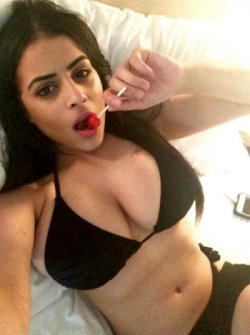 Brunette woman in black lingerie lying on a bed, holding a lollipop to her lips, taking a selfie.