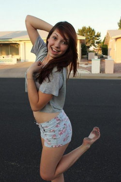 A smiling woman with long brown hair poses barefoot in a driveway, wearing a gray crop top and floral shorts.