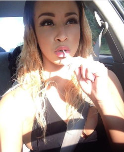 A woman with long ombre hair sits in a car, wearing a black top and holding her finger to her lips in a playful pose.
