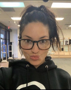 Woman with dark hair in a messy bun, wearing black glasses and a black hoodie, making a playful face in a store setting.