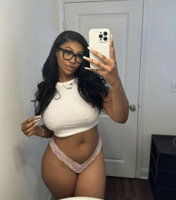 A curvy woman with long dark hair and glasses takes a mirror selfie in a white crop top and pink lace thong, holding a