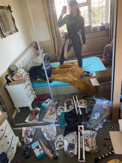 A woman in a black long-sleeve top and patterned pants takes a mirror selfie in a cluttered bedroom filled with adult