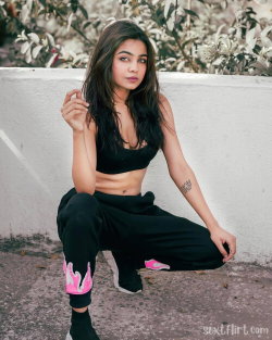 A young woman with long dark hair sits casually against a white wall, wearing a black crop top and sweatpants with pink