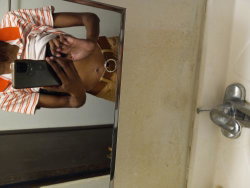 A person with dark skin takes a mirror selfie in a bathroom, wearing an orange and white striped shirt and a brown