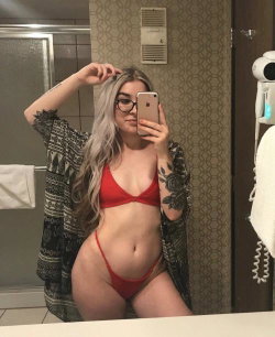 A woman with long gray hair and glasses takes a mirror selfie in a red bikini, wearing a patterned robe open over her