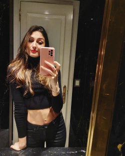 A woman with long, wavy hair and red lipstick takes a selfie in a black crop top and jeans.