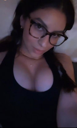 Woman with dark hair and glasses wearing a black halter top, looking at the camera in a dimly lit indoor setting.