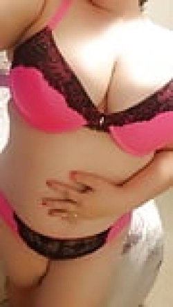 Woman in pink and black lace lingerie, curvy body type, posing indoors with hand on hip.
