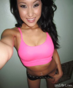 Asian woman with long dark hair in a bright pink crop top and black patterned bottoms, taking a selfie indoors.