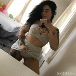 A woman with dark hair and multiple tattoos takes a mirror selfie in a lace crop top and shorts, standing in a bedroom