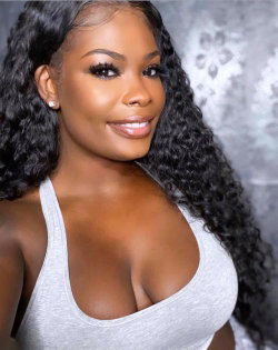 A woman with long curly black hair and a gray tank top poses against a patterned gray background.