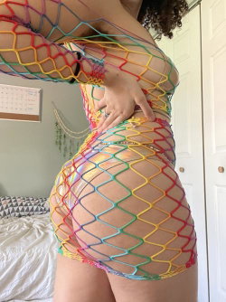 Curvy woman in colorful rainbow mesh bodysuit posing in bedroom, hand on hip, curly hair visible.