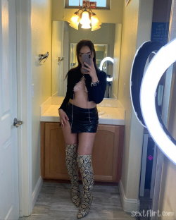 A woman with dark hair takes a mirror selfie in a bathroom, wearing a black crop top, shiny mini skirt, and snake-print