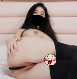 A woman with long dark hair lies on a bed, wearing black underwear and a face mask, her bare breasts visible.