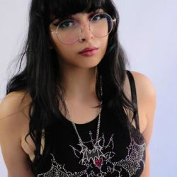 A woman with long black hair and round glasses wears a black embellished tank top, posing against a plain light