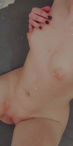 Close-up of a person's bare torso lying on a dark surface, showing a navel piercing and black-painted nails.