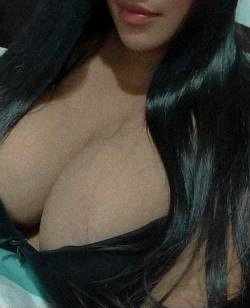 Brunette woman with long dark hair, wearing a black top that exposes her chest, in a close-up selfie-style shot.