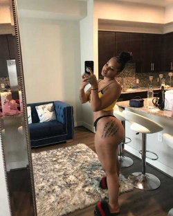 Curvy woman in yellow top and black thong takes mirror selfie in modern kitchen-living space, showing tattooed buttocks.