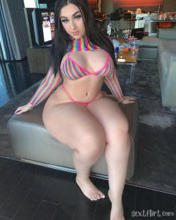 A curvy woman with long dark hair wears a colorful striped top and pink thong, sitting barefoot on an ottoman in a