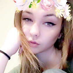 A young woman with blonde hair and a floral crown rests her chin on her hand, looking softly at the camera in a