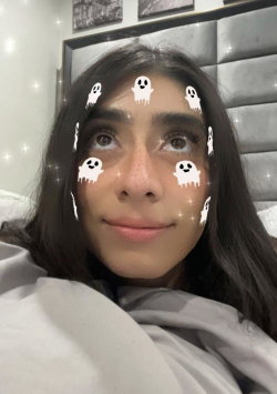 A young woman with long dark hair lies in bed under a gray blanket, using a ghost-themed Snapchat filter with sparkles.
