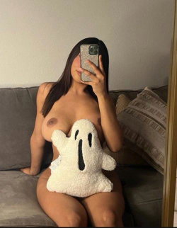 Curvy woman with long dark hair takes mirror selfie topless, holding phone, sitting on gray couch with plush ghost toy