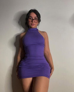 Woman with dark hair and glasses wearing a tight purple halter mini dress, standing against a plain wall.