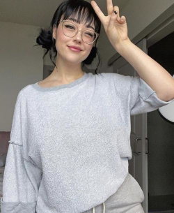 A woman with dark hair and glasses wears a loose gray sweatshirt, making a peace sign in a casual indoor setting.