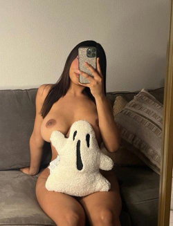 Brunette woman with curvy figure takes mirror selfie on gray couch, covering lower body with ghost plushie, bare