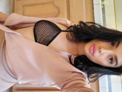 Brunette woman with long dark hair wearing a black mesh bra and open pink robe, taking a close-up selfie indoors.