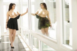 A curvy woman with dark hair stands topless in a black skirt and heels, touching a mirrored wall in a bright, modern
