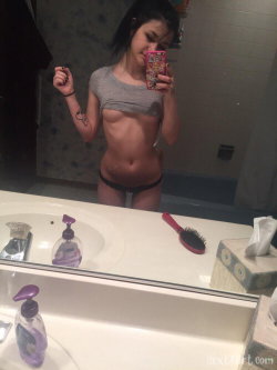 A young woman with dark hair takes a mirror selfie in a bathroom, wearing a gray crop top and black thong underwear.