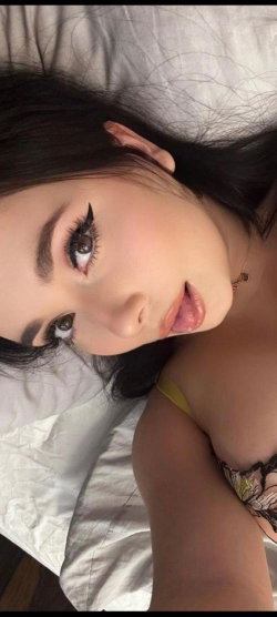 A woman with dark hair and makeup lies on a bed, wearing a yellow-strapped lace top, looking directly at the camera.