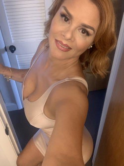 A woman with shoulder-length blonde hair poses in a beige bodysuit, smiling at the camera in a doorway setting.