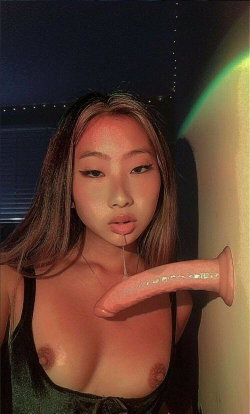 Asian woman with long hair, wearing a dark top, holding a sex toy near her bare chest in a dimly lit room.