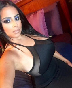 A woman with long dark hair and bold eye makeup poses in a black sheer top on a bed with purple pillows.