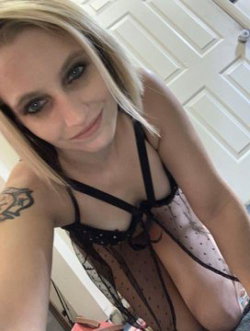 Blonde woman with tattoos wearing a black lace top, taking a selfie indoors near a white door.