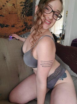 A smiling woman with curly hair and glasses wears a gray bra and lace underwear, sitting on a couch with visible